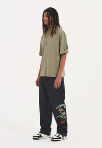 Green cotton t-shirt with a loose fit, paired with black denim pants featuring colorful dragon embroidery on the left leg and white sneakers.