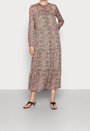 Kobieta ubrana w długą, sięgającą kostek sukienkę w kwiaty z warstwową spódnicą oraz czarne sandały na niskim obcasie, stojąca na tle jednolitego tła.