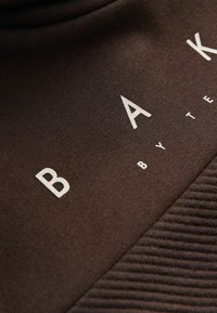 Tmavohnedá tkanina s rebrovanou textúrou a výrazným bielym nápisom "BAK" s "by TE" pod ním. Hladký povrch, detailný pohľad.