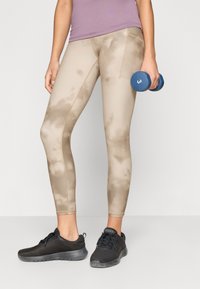 Μπεζ κολάν με σχέδιο tie-dye και ραφές σε αντίθεση, συνδυασμένα με μαύρα αθλητικά παπούτσια από δίχτυ. Το άτομο κρατά έναν μπλε αλτήρα.