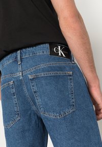 Jeans de mezclilla azul con un corte ajustado, que cuentan con una etiqueta negra de Calvin Klein en la cinturilla y costuras naranjas en contraste en los bolsillos.