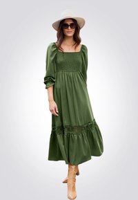 Abito lungo verde con scollatura quadrata, corpino arricciato, maniche a sbuffo, orlo in pizzo e tessuto morbido. Indossato con stivali beige al ginocchio.