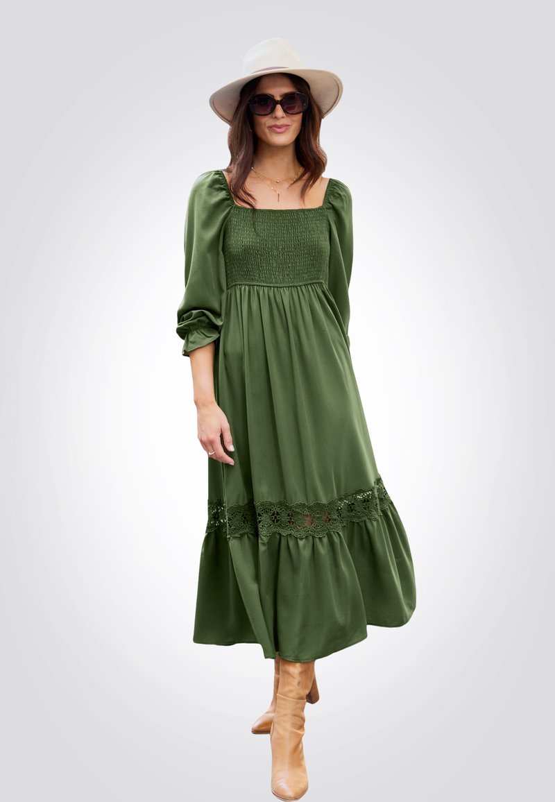 Abito lungo verde con scollatura quadrata, corpino arricciato, maniche a sbuffo, orlo in pizzo e tessuto morbido. Indossato con stivali beige al ginocchio.