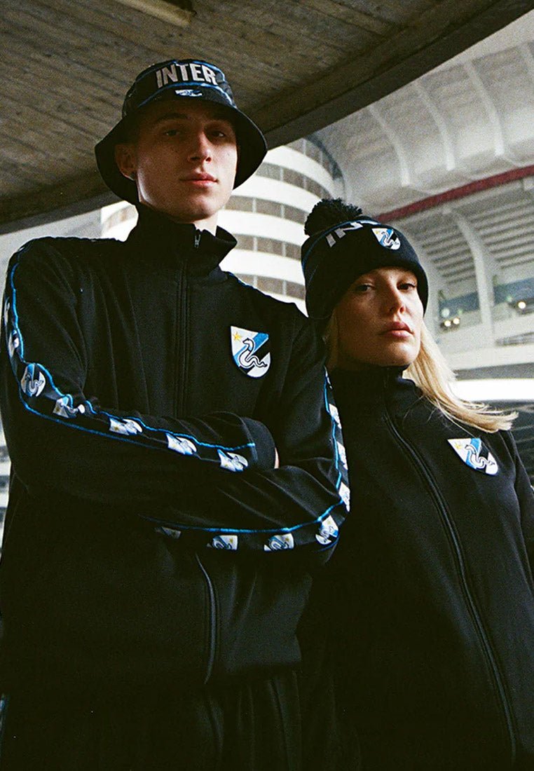 Zwarte zip-up jassen met blauwe accenten en logo's, gedragen met bijpassende zwarte beanies en bucket hats, met een pompon detail.