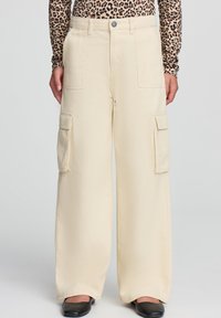 Szerokie spodnie cargo w kremowej bawełnie z bocznymi kieszeniami, zapięciem na guzik i gładką strukturą. Noszone z wzorzystą bluzką z długim rękawem.
