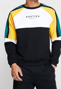 Svart sweatshirt med flerfärgade raglanärmar i gult, teal och vitt. Har texten "KAOTIKO since 1999" på bröstet. Bekväm passform.