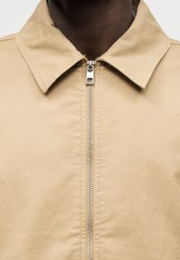 Μπεζ μπουφάν με φερμουάρ και μυτερό γιακά, φορεμένο από άτομο με σκουρόχρωμο δέρμα, εμφανιζόμενο από τον λαιμό μέχρι το στήθος.