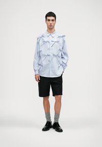 Camisa de mangas longas azul-claro com vários laços de tecido, combinada com calções pretos, meias cinzas e sapatos pretos. Design simples e estruturado.