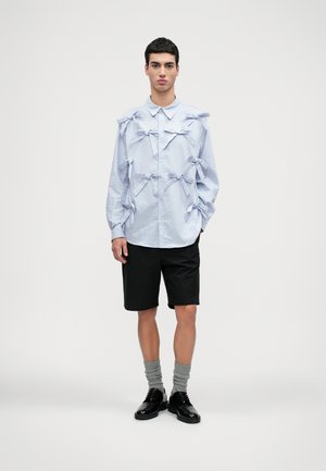 Helles blaues Langarmhemd mit mehreren stofflichen Schleifen, kombiniert mit schwarzen Shorts, grauen Socken und schwarzen Schuhen. Schlichtes, strukturiertes Design.