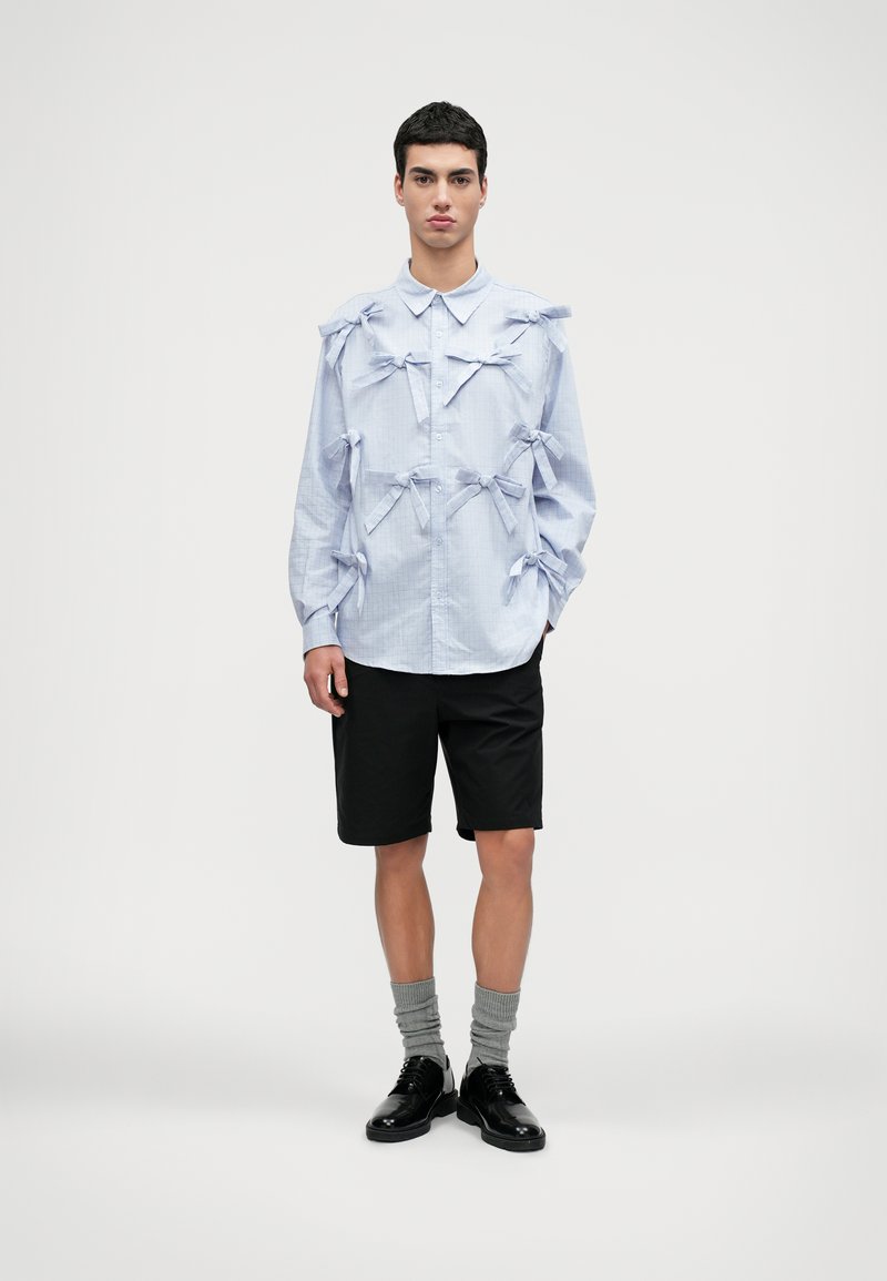 Camisa de mangas longas azul-claro com vários laços de tecido, combinada com calções pretos, meias cinzas e sapatos pretos. Design simples e estruturado.