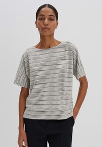 White short-sleeve top featuring a black zigzag pattern, soft fabric, wide neckline, and relaxed fit, paired with dark pants.