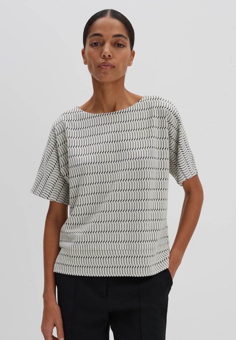 White short-sleeve top featuring a black zigzag pattern, soft fabric, wide neckline, and relaxed fit, paired with dark pants.