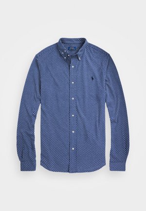 Chemise bleue à manches longues, boutonnée, à pois blancs avec un petit logo brodé sur la poitrine gauche.