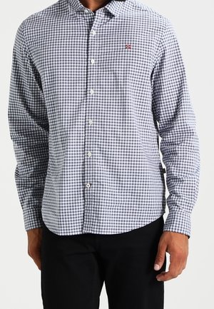 Mann trägt ein grau-weiß kariertes, langärmeliges Hemd mit Knöpfen und einem kleinen gestickten Logo auf der Brust, kombiniert mit schwarzer Hose.