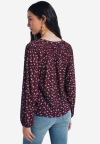 Blusa bordeaux a fiori con maniche lunghe a sbuffo e collo smock, caratterizzata da un delicato motivo floreale bianco e blu.