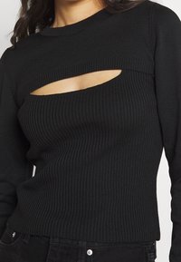 Haut en maille côtelée noir avec un design découpé au niveau de la poitrine, des manches longues et une finition texturée. Silhouette simple et ajustée.