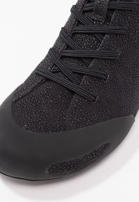 Black athletic shoe with a textured upper, lace-up closure, and a smooth rubber toe cap. Features a sleek, modern design with subtle detailing.