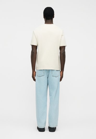 Άνδρας με την πλάτη γυρισμένη φοράει απλό, λευκό κοντομάνικο μπλουζάκι, ανοιχτό μπλε τζιν παντελόνι άνετης γραμμής και μαύρα παπούτσια σε λευκό φόντο.