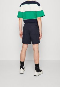 Camisa de manga corta en bloques de colores verde, marino y crema; combinada con pantalones cortos marino. El modelo lleva calcetines negros y zapatillas blancas.