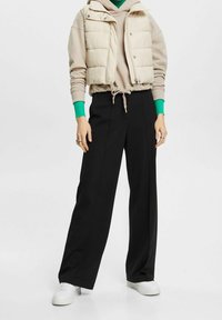 Beige pufferväst över en beige huvtröja med gröna detaljer, kombinerat med högmidjade svarta vida byxor och vita sneakers.