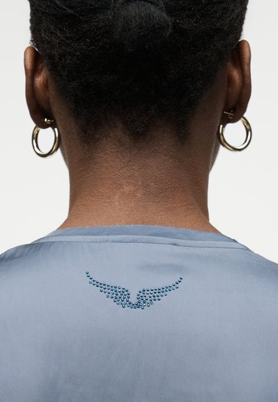Πλάτη ατόμου με πλεξούδες που φορά μπλε τοπ με μικρό σχέδιο μπλε φτερών από στρας στη λαιμόκοψη και χρυσούς κρίκους σκουλαρίκια.