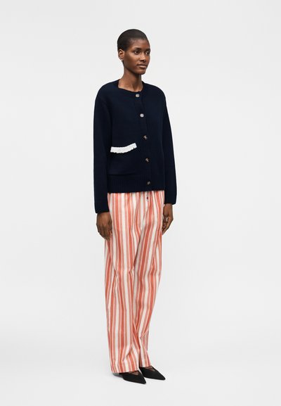 Γυναίκα που στέκεται, φοράει μαύρη ζακέτα με κουμπιά και άσπρη επένδυση στην τσέπη, ριγέ πορτοκαλί και λευκά παντελόνια με φαρδιά πόδια και μαύρα μυτερά παπούτσια.