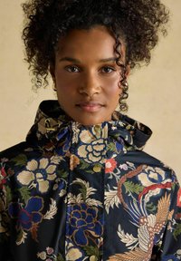 Tânără cu păr creț purtând o jachetă cu glugă, cu model floral închis la culoare și nasturi aurii, privind înainte pe un fundal neutru.