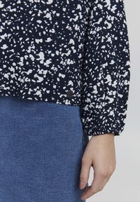 Blusa blu navy con motivo astratto bianco, maniche lunghe con polsini elasticizzati, abbinata a una gonna di jeans blu. Dettaglio in metallo all'orlo.