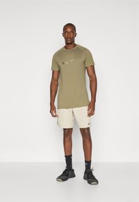 Olivengrünes, kurzärmeliges Sport-T-Shirt mit metallic Nike-Logo, kombiniert mit beigen Shorts und schwarzen Sneakern, auf einem weißen Hintergrund.