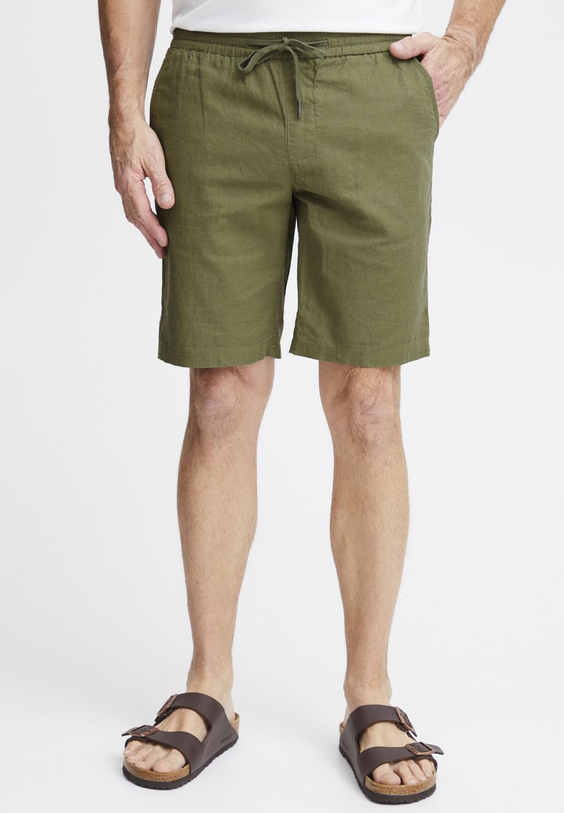 Pantaloni corti in lino verde oliva con vita elasticizzata, chiusura con cordoncino e tasche laterali, abbinati a sandali regolabili marroni.