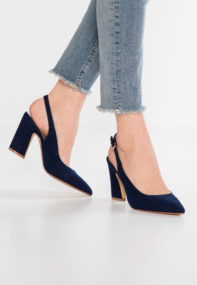 Femme portant des escarpins slingback en daim bleu et un jean bleu clair à ourlet effiloché, debout avec un pied légèrement relevé.