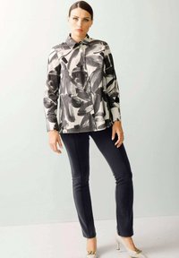 Langärmlige, knöpfbare Bluse im schwarz-weißen abstrakten Muster, kombiniert mit eng anliegenden schwarzen Hosen und beigen High Heels. Glatte Stoffoberfläche.