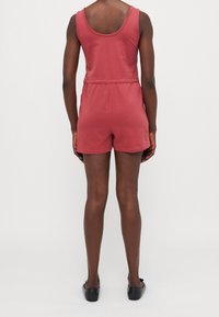 Femme à la peau foncée portant une combinaison rouge sans manches et des chaussures plates noires, debout de dos sur un fond blanc uni.