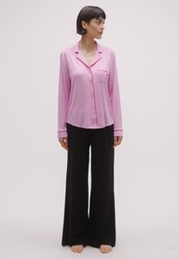 Set de pijamale din satin roz, cu o cămașă cu nasturi și buzunar, decorată cu margini roșii, asortată cu pantaloni negri cu picioare largi. Textură netedă evidentă.