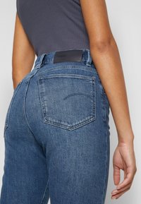 Jeans taille haute bleu foncé avec une texture lisse, deux poches arrière avec couture et un patch en cuir noir à la taille.