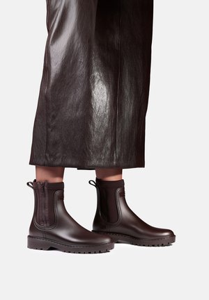 Bottines marron avec fermetures éclair latérales portées avec une longue jupe en cuir marron foncé sur un fond gris clair.