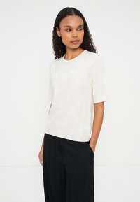 Kreklu ar īsām piedurknēm, gaišā krāsā, ar ziedu rakstu, izgatavots no mīksta auduma. Apaļš kakla izgriezums un brīvs piegriezums, apvienots ar tumšām platām biksēm.