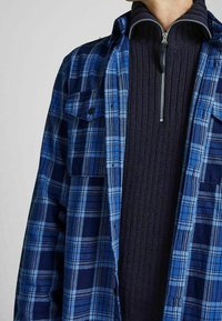 Koszula w kratę flanelowa w kolorze niebieskim i granatowym, z dwiema kieszeniami na piersi oraz ciemnym swetrem z zamkiem błyskawicznym na dole, z prążkowanym wzorem.