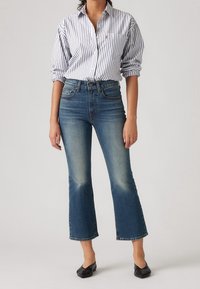 Jean évasé bleu avec décoloration aux genoux, taille haute et design à cinq poches, associé à une chemise boutonnée blanche à rayures bleu marine.