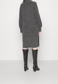 Robe pull tricotée gris foncé avec un col montant, des manches longues, des fentes latérales et un ourlet côtelé ; associée à des bottes en cuir noir jusqu'aux genoux.