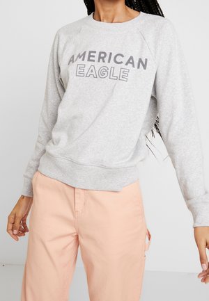 Sudadera gris con la inscripción "AMERICAN EAGLE" en gris oscuro. Presenta puños y dobladillo acanalados y un ajuste cómodo. Combinada con pantalones rosa claro.