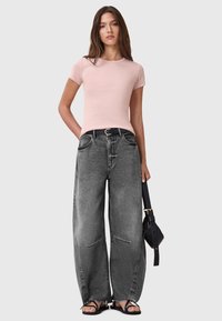 Lichtroze slim-fit t-shirt, grijze wijde spijkerbroek met versleten zoom, zwarte riem, zwarte handtas en platte sandalen. Eenvoudige, casual outfit.