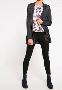 Dlouhý kardigan s černobílým vzorem přes květinovou blůzu, soutředěné s černými skinny kalhotami a kotníkovými botami. Texturovaná tkanina, uvolněný střih.