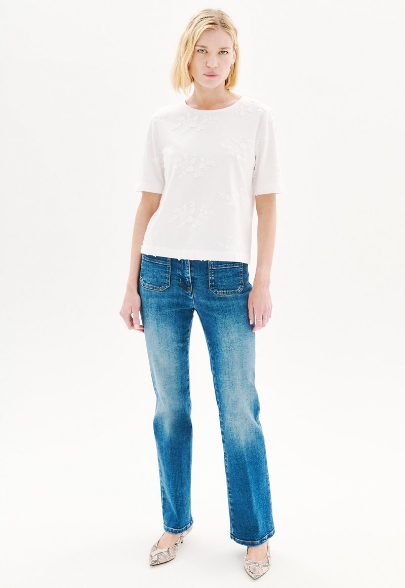 Camisa blanca de manga corta con bordado floral, combinada con unos jeans rectos azul desgastado y tacones de estampado de serpiente. Diseño simple y limpio.