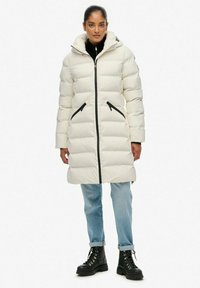 Witte donsjas met capuchon, voorzien van zwarte ritsen en zijzakken. De gladde textuur vormt een mooie combinatie met relaxte blauwe jeans en zwarte laarzen.