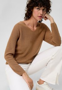 Pull en maille côtelée marron avec un col en V, assorti à un pantalon blanc à contraste élevé et des baskets légères, mettant en avant une coupe décontractée et une texture.