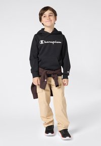 Un giovane ragazzo con indosso un hoodie nero della Champion, pantaloni cargo beige, scarpe da ginnastica nere e un maglione marrone legato in vita, in piedi su uno sfondo bianco.