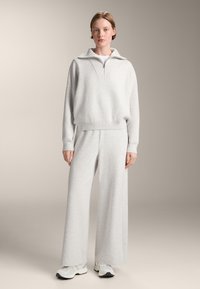 Grauer, übergroßer Sweatshirt mit hohem Kragen und Halbreißverschluss, kombiniert mit weiten grauen Hosen. Das Material wirkt weich mit einer Strickstruktur.