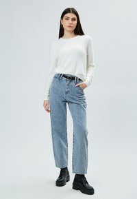 Maglione bianco a trama, jeans azzurri a vita alta, cintura nera con fibbia tonda, stivali neri chunky, in pose di standing su uno sfondo neutro.