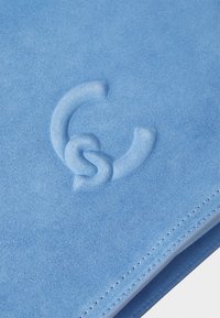 Matériau en suède bleu avec une texture douce, présentant un détail de logo en relief. Un surfilage soigné le long du bord ajoute à son design.